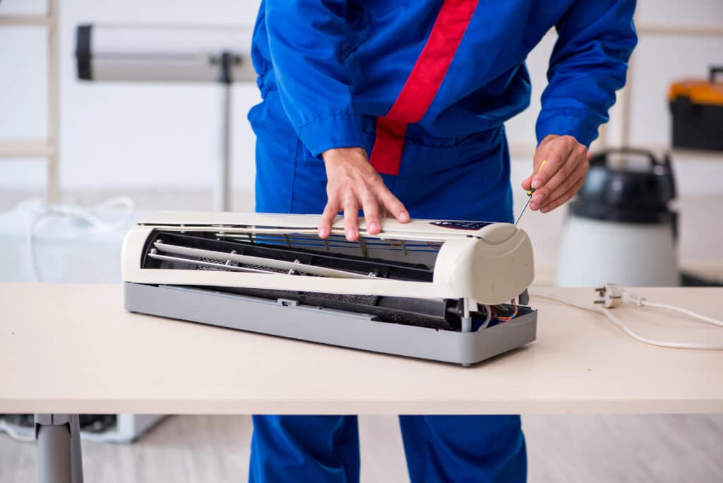 Réparation de climatiseur : service par un technicien CVC qualifié Technicien CVC en bleu et rouge démontant un climatiseur mural sur table avec un tournevis. Réparation et entretien.